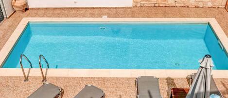 A rectangular pool with clear blue water, surrounded by lounge chairs and a covered umbrella.