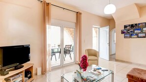 A living room with a TV, bookshelves, a glass coffee table, a chair, and a sofa. - Villa in Kalamitsi Near White Mountains (Apokoronas)