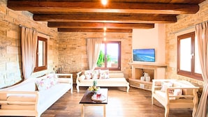 Una sala de estar acogedora con paredes de piedra, vigas de madera en el techo y una chimenea. Hay dos sofás, una mesa de centro y un televisor montado en la pared.