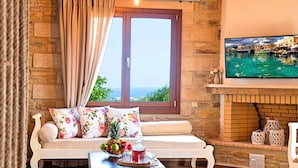 Una habitación acogedora con pared de piedra, suelo de madera, sofá blanco con cojines florales, una pequeña mesa con fruta y bebidas, y un televisor que muestra una escena costera.