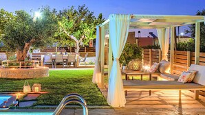 Un área junto a la piscina con terraza de madera, un espacio de descanso cubierto con cojines y un césped bien cuidado.
