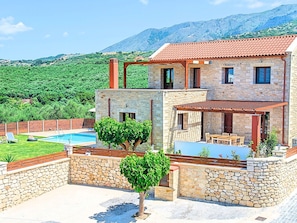 A two-story stone house with a swimming pool, outdoor seating area, and a view of green hills. - Luxurious Villa With Swimming Pool in Kavallos Greece (Apokoronas)
