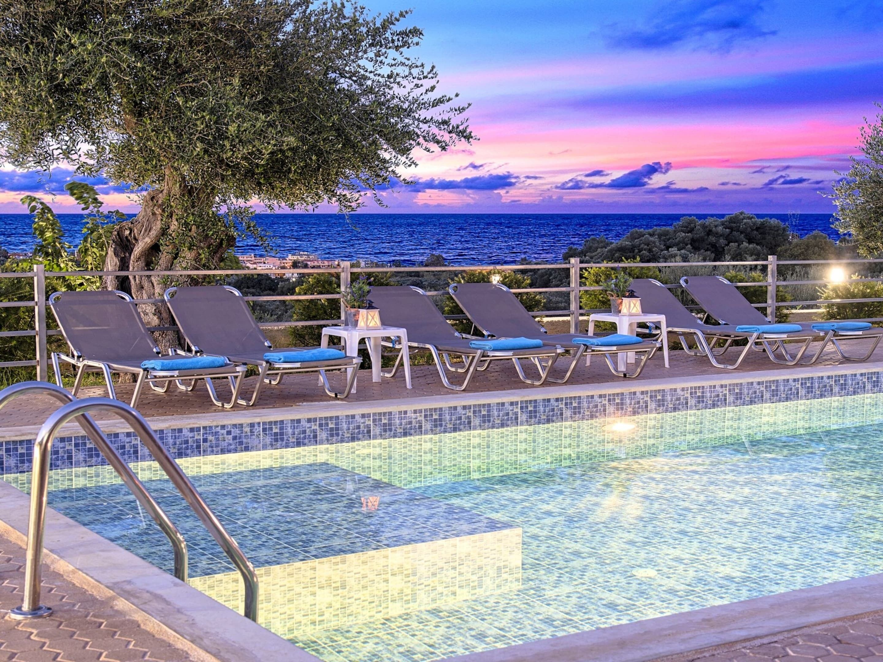 A poolside area with lounge chairs and a view of the sea at sunset.