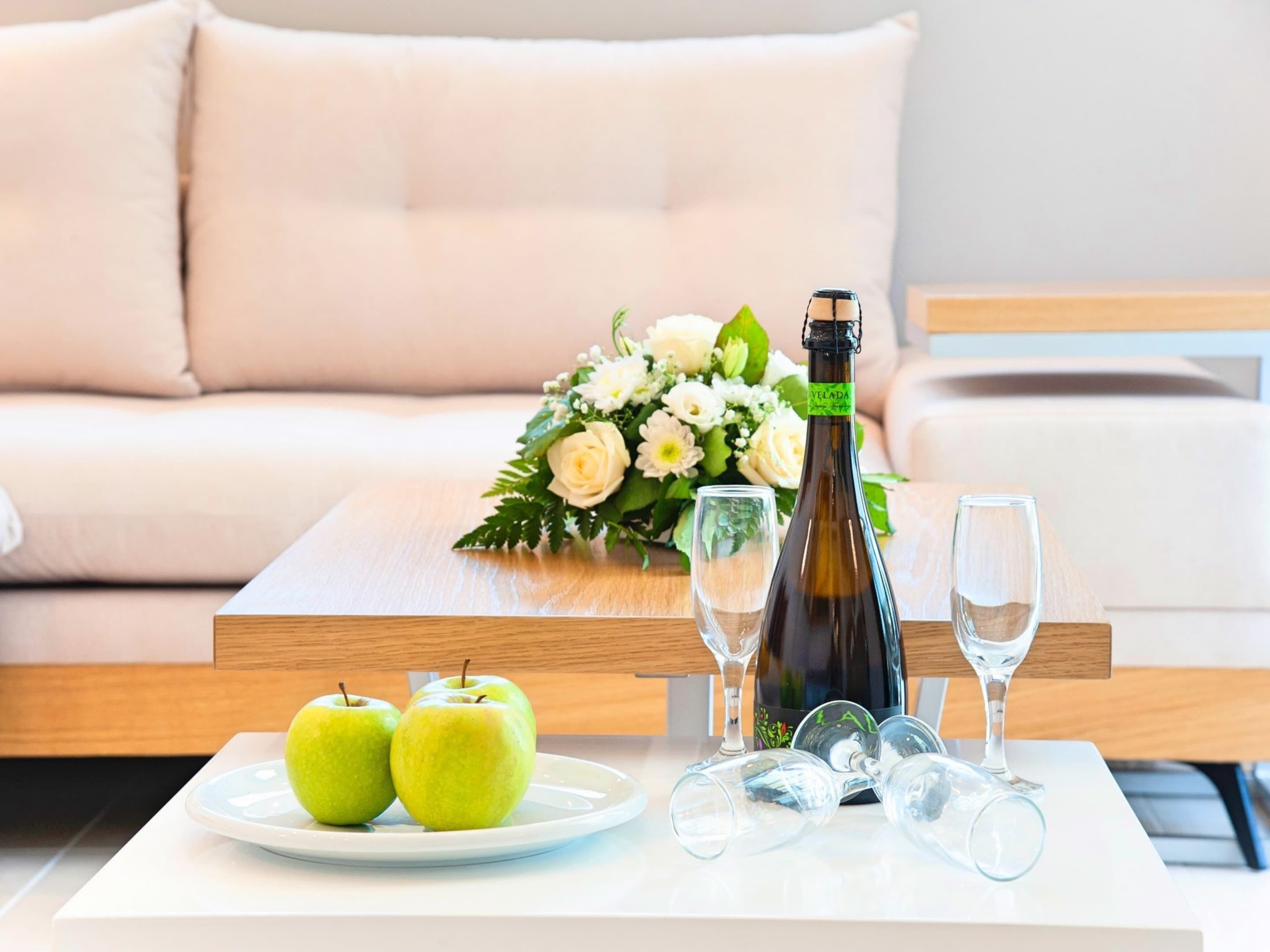 A table with a bottle of champagne, two glasses, a plate with two apples, and a bouquet of flowers.