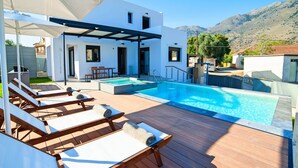 A modern outdoor pool area with wooden loungers and a covered seating area, surrounded by a white building and mountains in the background. - 2 new Villas With Heated Pool, Rent Separately or Together at Kournas Lake (Apokoronas)