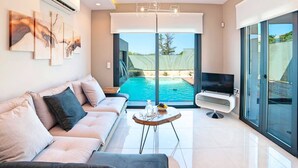 A modern living room with a sofa, a coffee table, a TV, and a pool view. - 2 New Villas Next Each Other, Pool, Rent Separately or Together at Kournas Lake (Apokoronas)
