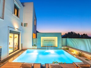 Ein moderner Poolbereich im Freien mit einem klaren, blauen Schwimmbecken, einer Holzterrasse und einem weißen Gebäude mit Glastür.