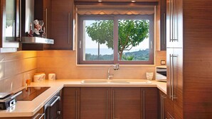 A modern kitchen with wooden cabinets, a built-in oven, a large window with a view of trees, and a stainless steel sink. - 2 Villas Next to Each Other with Pool & Sea View (Apokoronas)