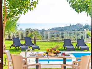 Un espace repas aménagé au bord de la piscine, avec une table pour deux, entouré de chaises longues, d’une piscine et offrant une vue sur la mer et les collines.