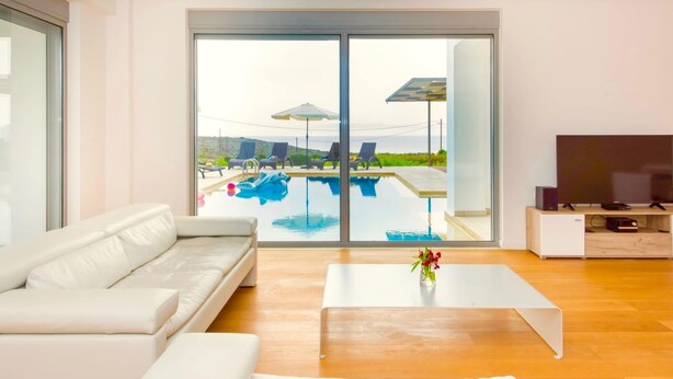 A modern living room with a white sofa, a coffee table, and a TV stand. A large sliding glass door leads to a pool area with lounge chairs and umbrellas. - Sparkling Villa in Acrotiri with Private Pool near Sea Beach (Chania)