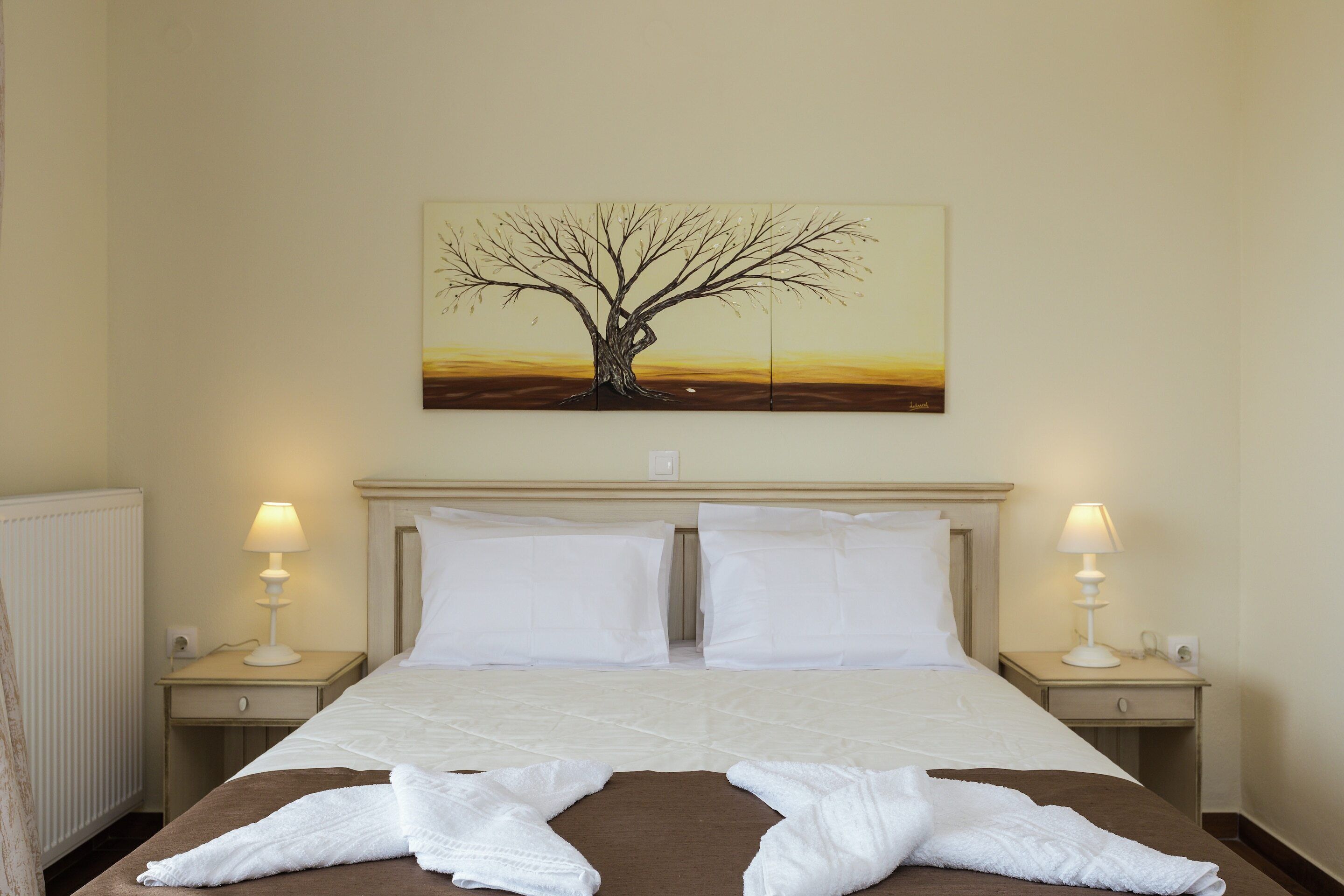 A hotel room with a large bed, two bedside tables with lamps, a painting of a tree on the wall, and a radiator on the left.