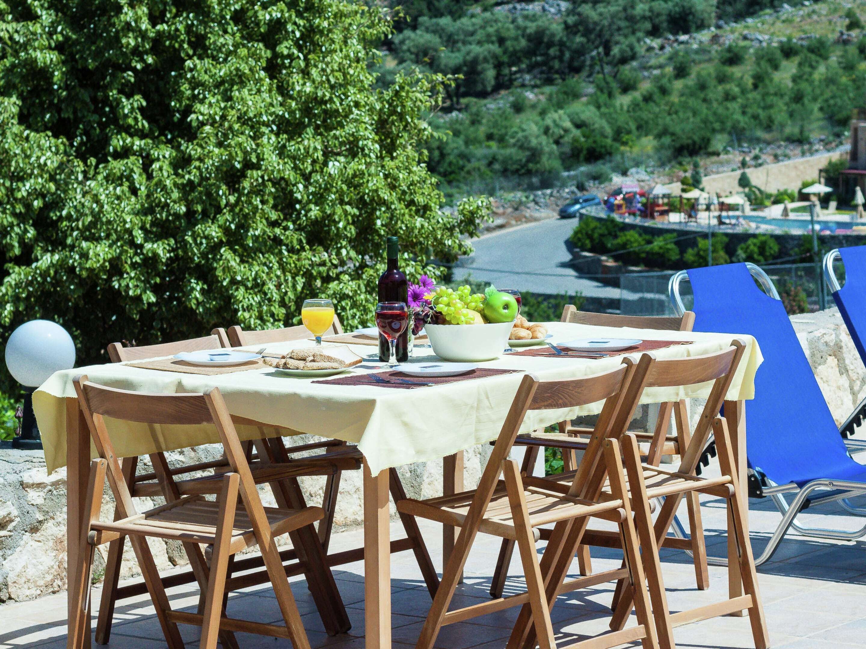 An outdoor dining area with a table set for two, surrounded by wooden chairs. A bowl of fruit and a bottle of wine are on the table.