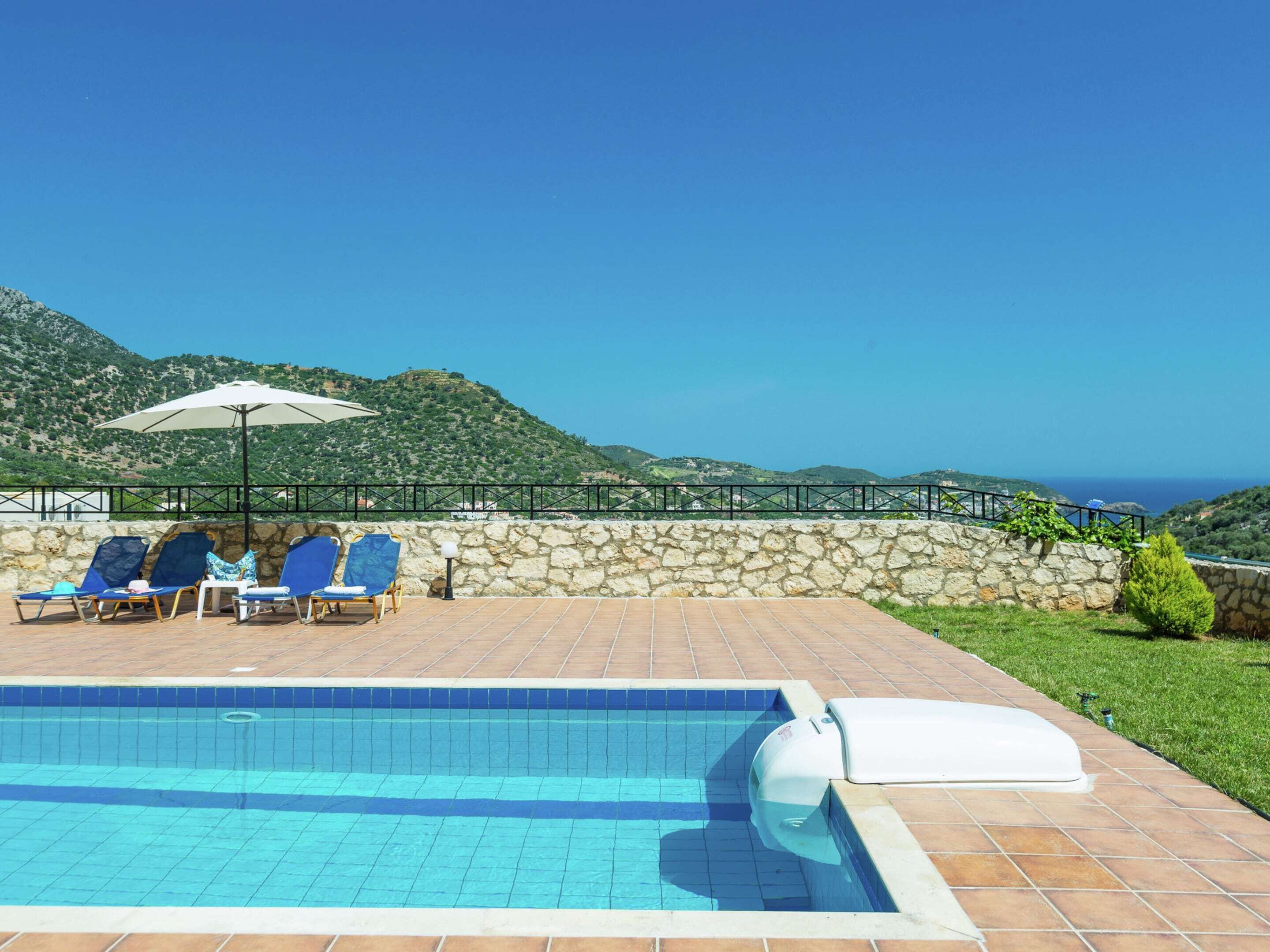 Zona de piscina con sillones de descanso y una sombrilla, rodeada de muros de piedra y vistas al mar y las colinas.