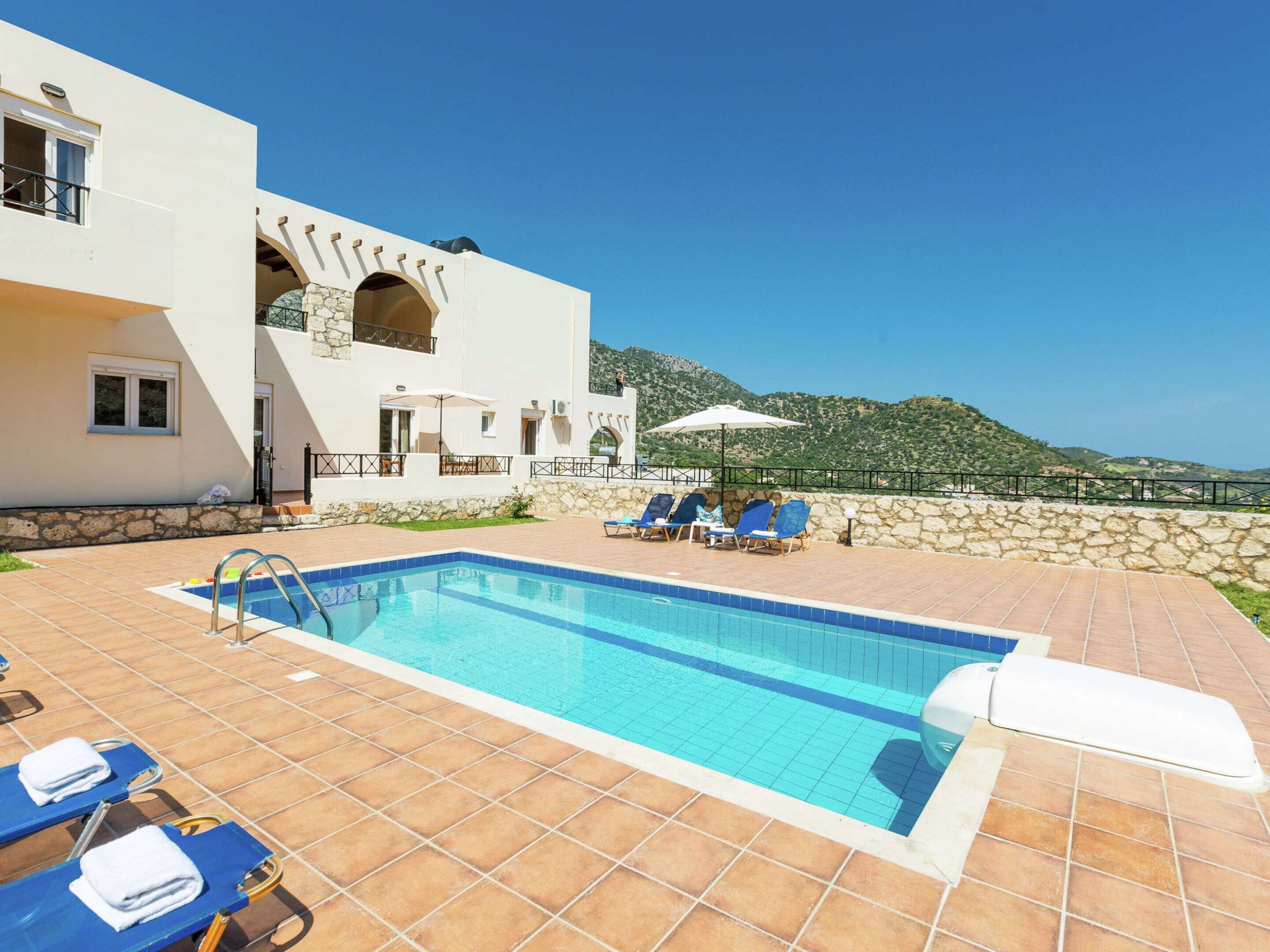 A pool area with blue lounge chairs, a clear sky, and a mountainous backdrop.