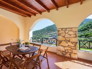 A wooden table with chairs and a book, set against a stone wall with a view of a green hillside and buildings.