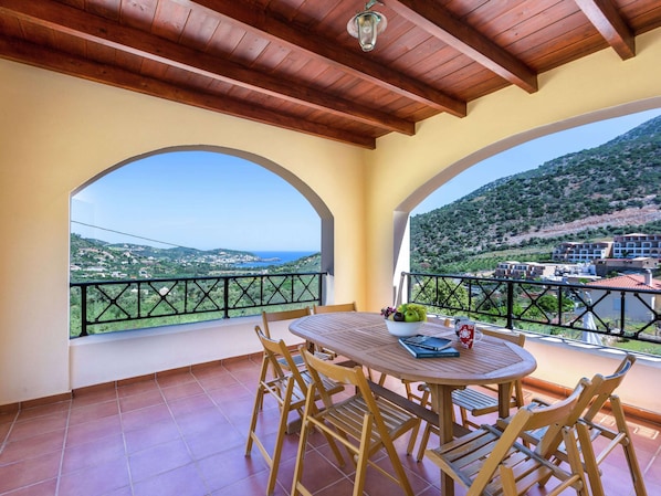 A balcony with a wooden table and chairs, a bowl of fruit, and a view of a coastal landscape with buildings and hills.