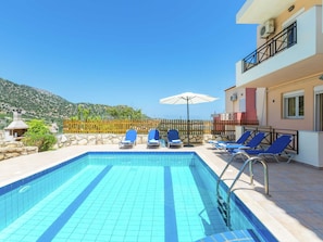 A swimming pool with blue tiles, surrounded by lounge chairs and umbrellas, with a mountain view in the background.