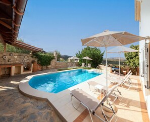 A swimming pool with lounge chairs and umbrellas, stone flooring, and a stone wall. - Villa in Crete With Pool & Olive Grove Views (Mylopotamos)