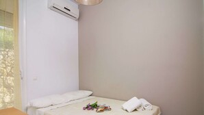 Een klein, net bed met witte lakens, een nachtkastje met een kop en een boek, en een airconditioner aan de muur.