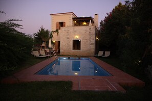 A rectangular swimming pool with a stone wall surrounding it, a small deck area with chairs, and a building with a balcony in the background.