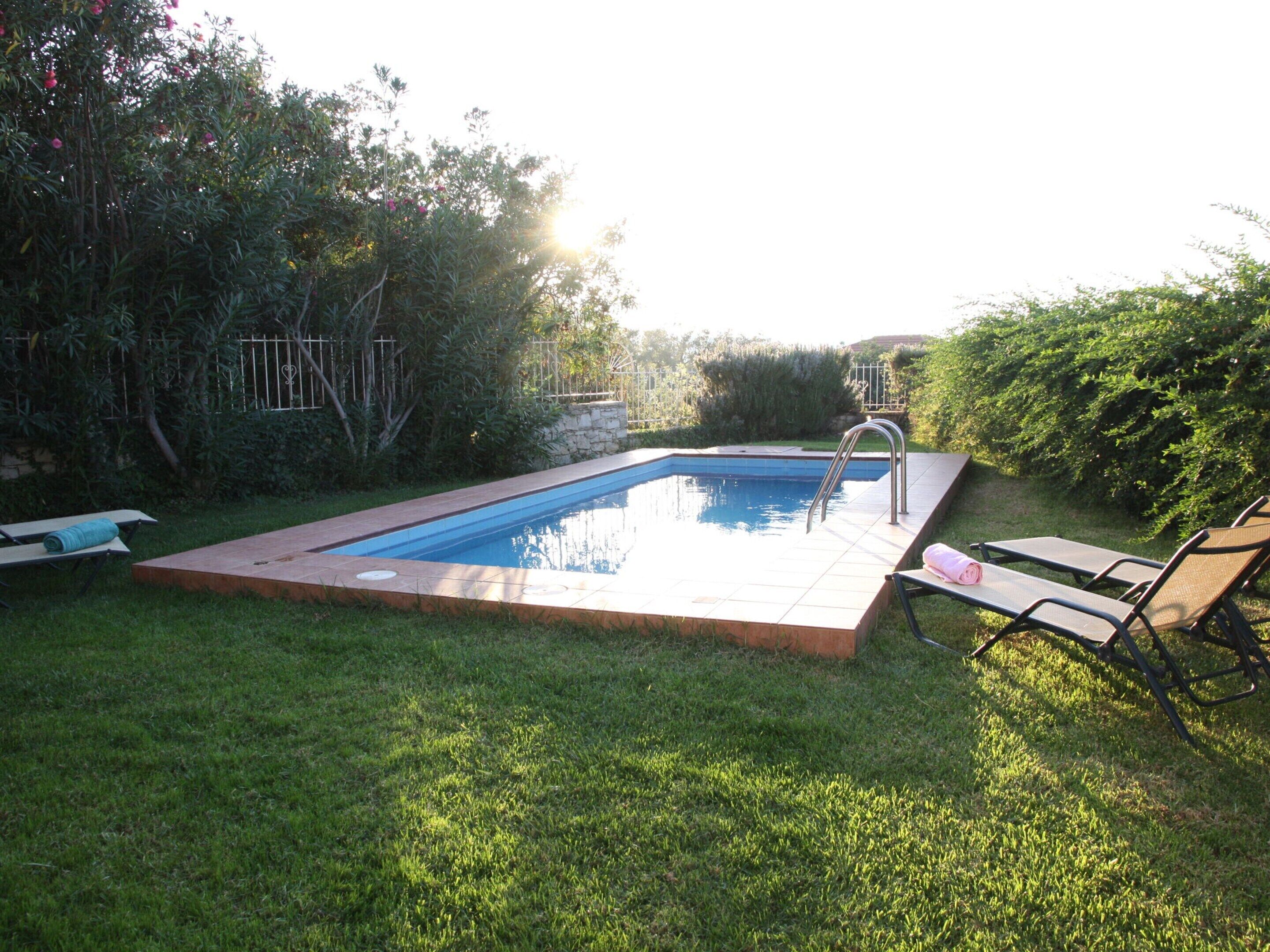 Ein rechteckiger Pool mit Fliesenrand, umgeben von Gras und Grünpflanzen. Zwei Liegestühle und ein Tisch mit einem Handtuch stehen bereit.