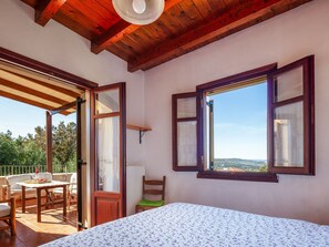 A bedroom with a bed, a window with a view, a wooden ceiling, and a balcony with outdoor furniture.