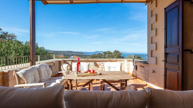 Eine Terrasse mit Holztisch, weiß gepolsterten Sitzmöglichkeiten und Blick auf das Meer und die Hügel.