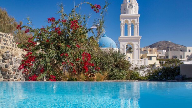 A clear blue pool with a white building featuring a clock tower and a blue dome in the background. - Extravagant Villa in Megalochori (Santorini)