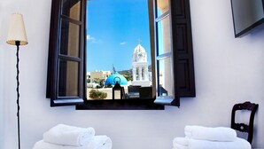 A hotel room with an open window revealing a view of a blue-domed building and a white church with a clock tower. There are two neatly folded white towels on a bed. - Extravagant Villa in Megalochori (Santorini)