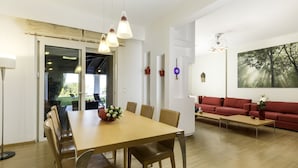 A modern living room with a dining area, featuring a wooden dining table, chairs, a red sofa, and a large window with a view of greenery. - Villa on Island With Private Pool (Rhodes)