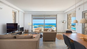 A modern living room with a flat-screen TV, beige sofas, a dining table, and a view of the sea through large windows. - Villa in Rhodes With Infinity Pool & Sea Views (Rhodes)