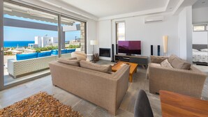 A modern living room with a sofa set, a coffee table, and a TV. There is a large window with a view of the sea and buildings. - Villa in Rhodes With Infinity Pool & Sea Views (Rhodes)