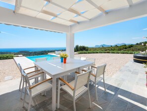 A covered outdoor dining area with a pool view, white furniture, and a clear sky. - Villa in Rhodes With Infinity Pool & Sea Views (Rhodes)