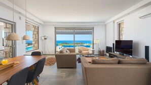 A modern living room with a wooden dining table, two sofas, a TV, and a large window with a view of the sea. - Villa in Rhodes With Infinity Pool & Sea Views (Rhodes)