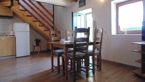 Uma área de refeições com mesa e cadeiras de madeira, uma cozinha com frigorífico branco, e uma escada de madeira.