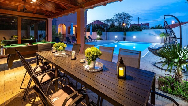 An outdoor pool area with a wooden deck, poolside chairs, and a dining table with chairs and centerpieces. - Luxurious Villa in Dalmatia Near Sea (Nin)
