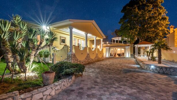 A well-lit building with a stone pathway leading to a covered entrance, surrounded by palm trees and a stone wall.