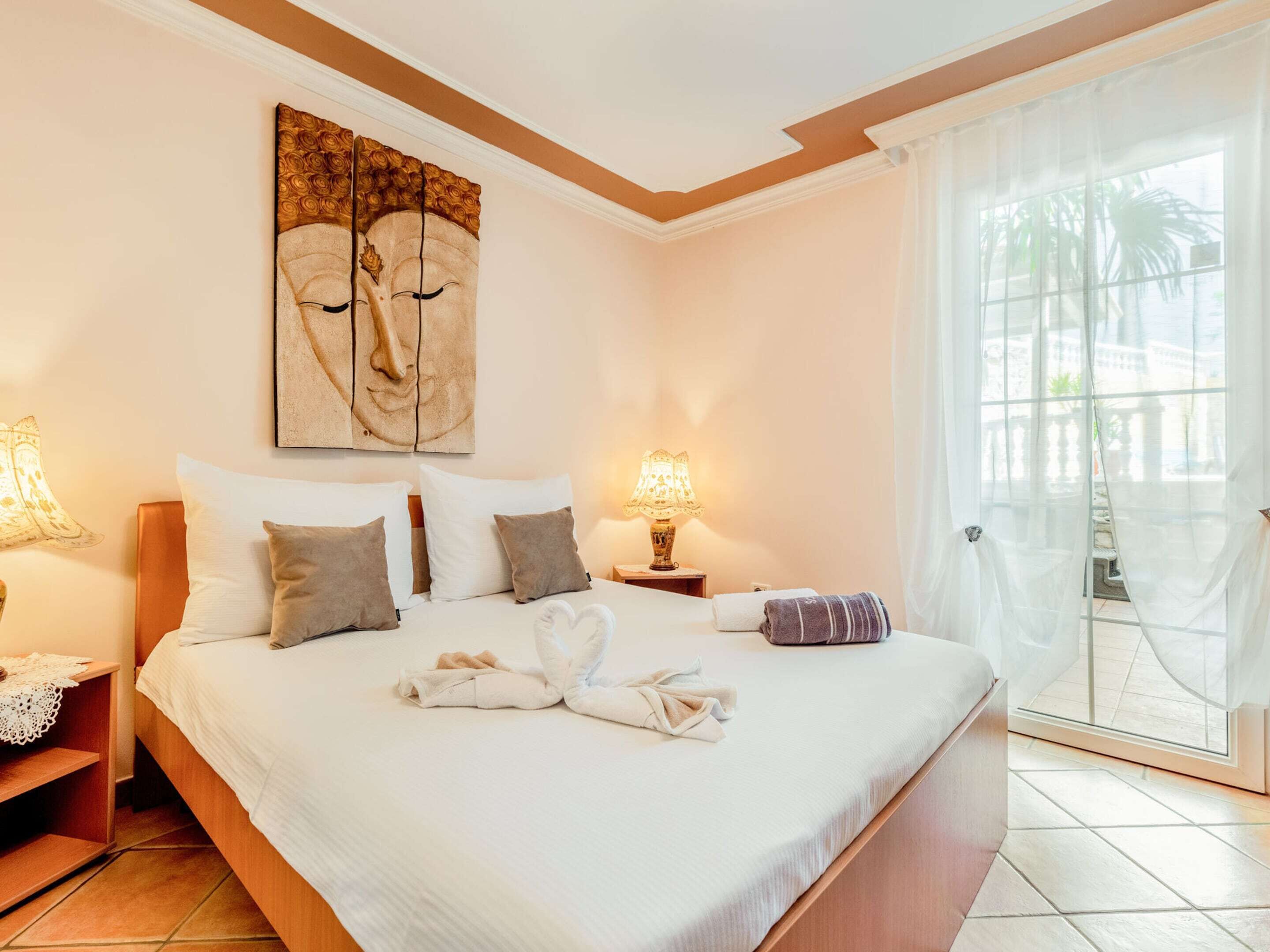 Habitación de hotel con una cama grande, dos lámparas de noche, un cuadro en la pared y vistas a palmeras a través de la ventana.