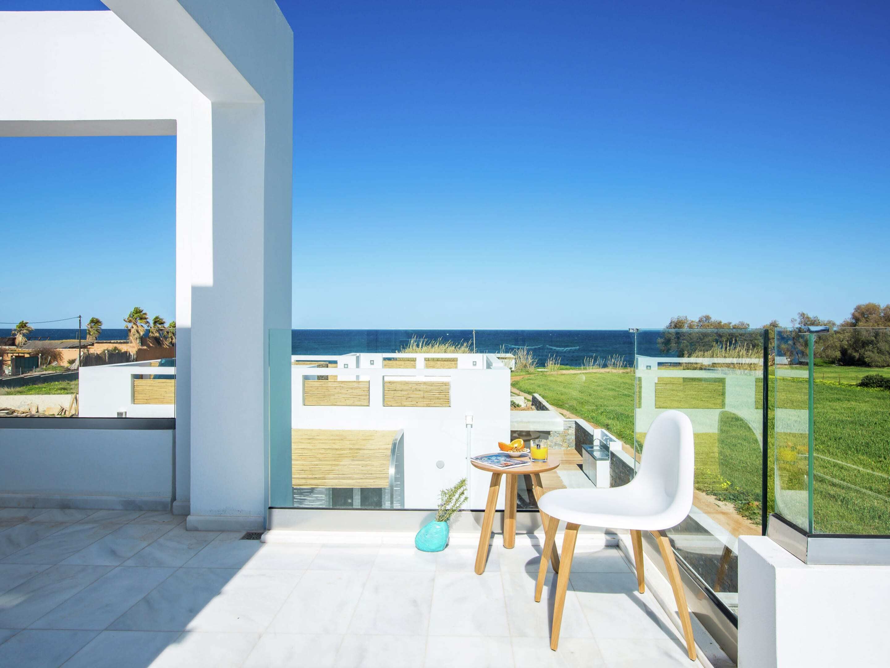 A balcony with a clear view of the sea, a white chair, a small table with a drink, and a glass railing.