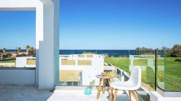 A balcony with a clear view of the sea, a white chair, a small table with a drink, and a glass railing.