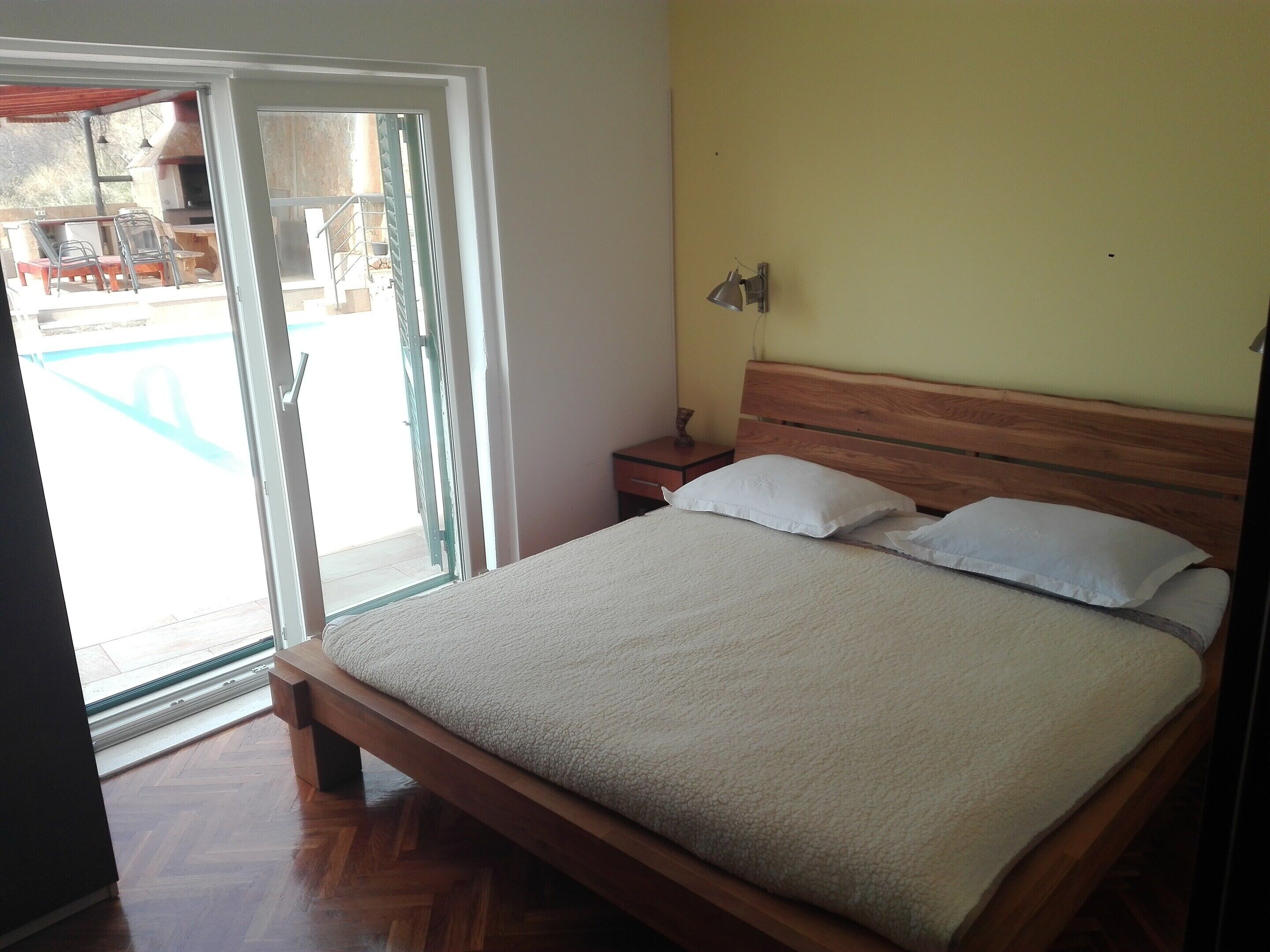 A bedroom with a wooden bed, a nightstand, a lamp, and a sliding glass door leading to an outdoor area with a barbecue.