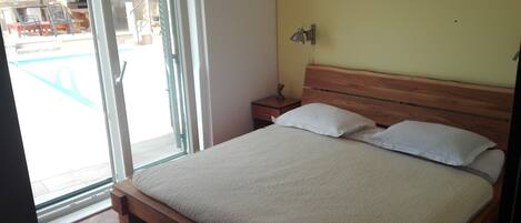 A bedroom with a wooden bed, a nightstand, a lamp, and a sliding glass door leading to an outdoor area with a barbecue.