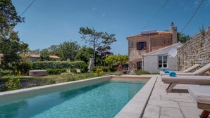 A rectangular swimming pool with clear water, surrounded by a stone deck. There are lounge chairs with towels, a stone wall, and a building with a tiled roof in the background. - Holiday Home in Osor Near Sandy Beach (Mali Losinj)