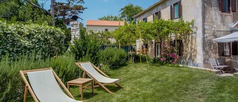 Una vila amb un jardí ben cuidat, dues gandules i un edifici de pedra amb teulada.