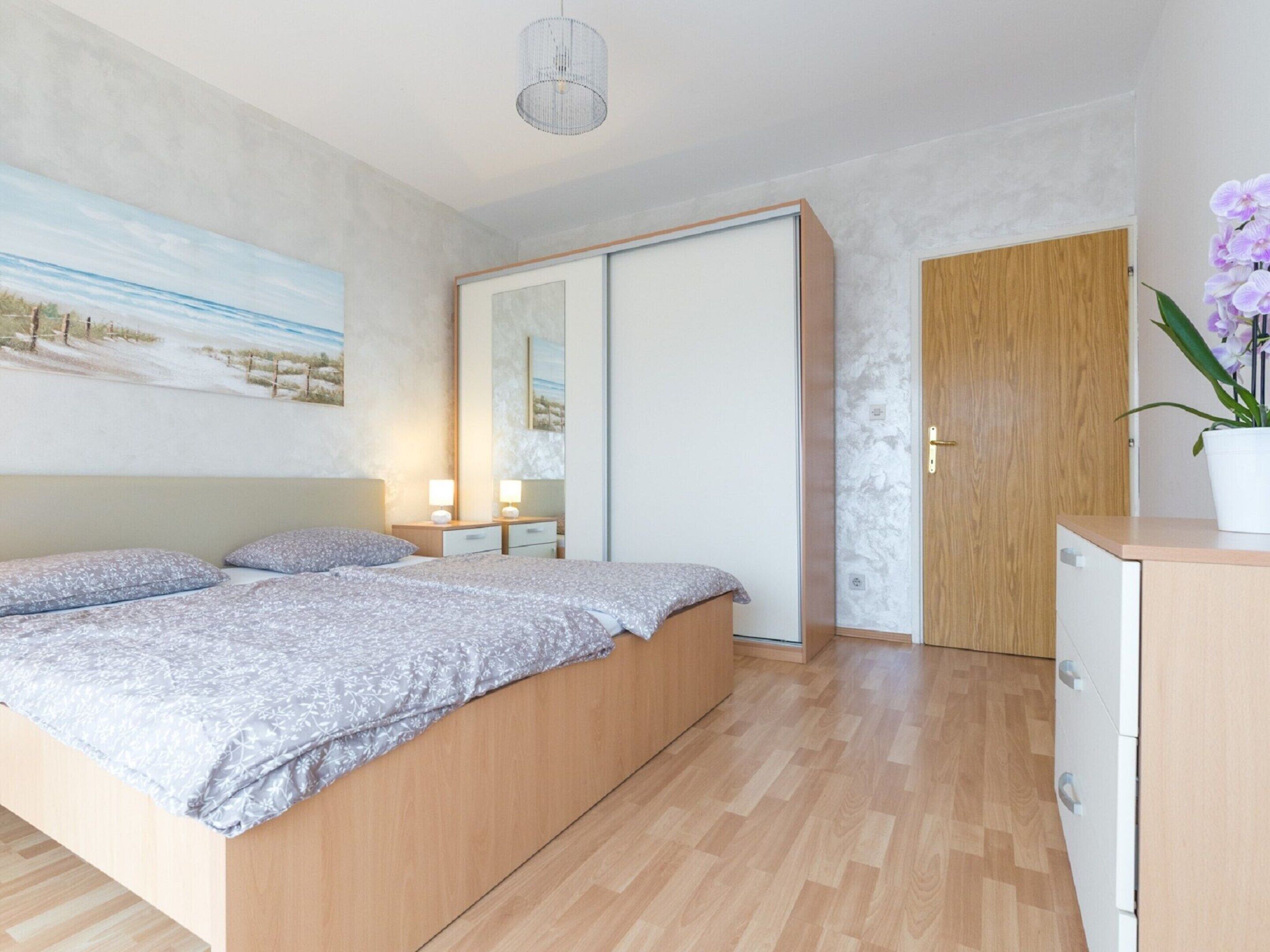 A modern bedroom with a wooden bed, a nightstand with lamps, a wardrobe, and a small cabinet.