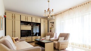 Una sala de estar con un sofá, dos sillones, una mesa de centro y un televisor. Hay armarios y una araña.
