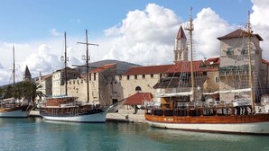 Un porto con diverse barche a vela ormeggiate, un edificio storico in pietra con un campanile, e un cielo azzurro limpido.
