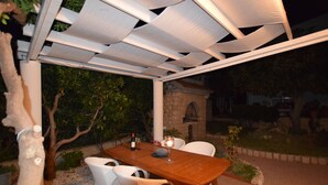 An outdoor dining area with a wooden table and white chairs, under a white pergola. - Comfortable Apartment With Balcony and Outdoor Kitchen (Pag)