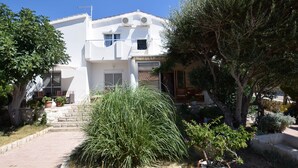 A white two-story house with a paved driveway, a small garden with various plants, and a wooden bench. - Comfortable Apartment With Balcony and Outdoor Kitchen (Pag)