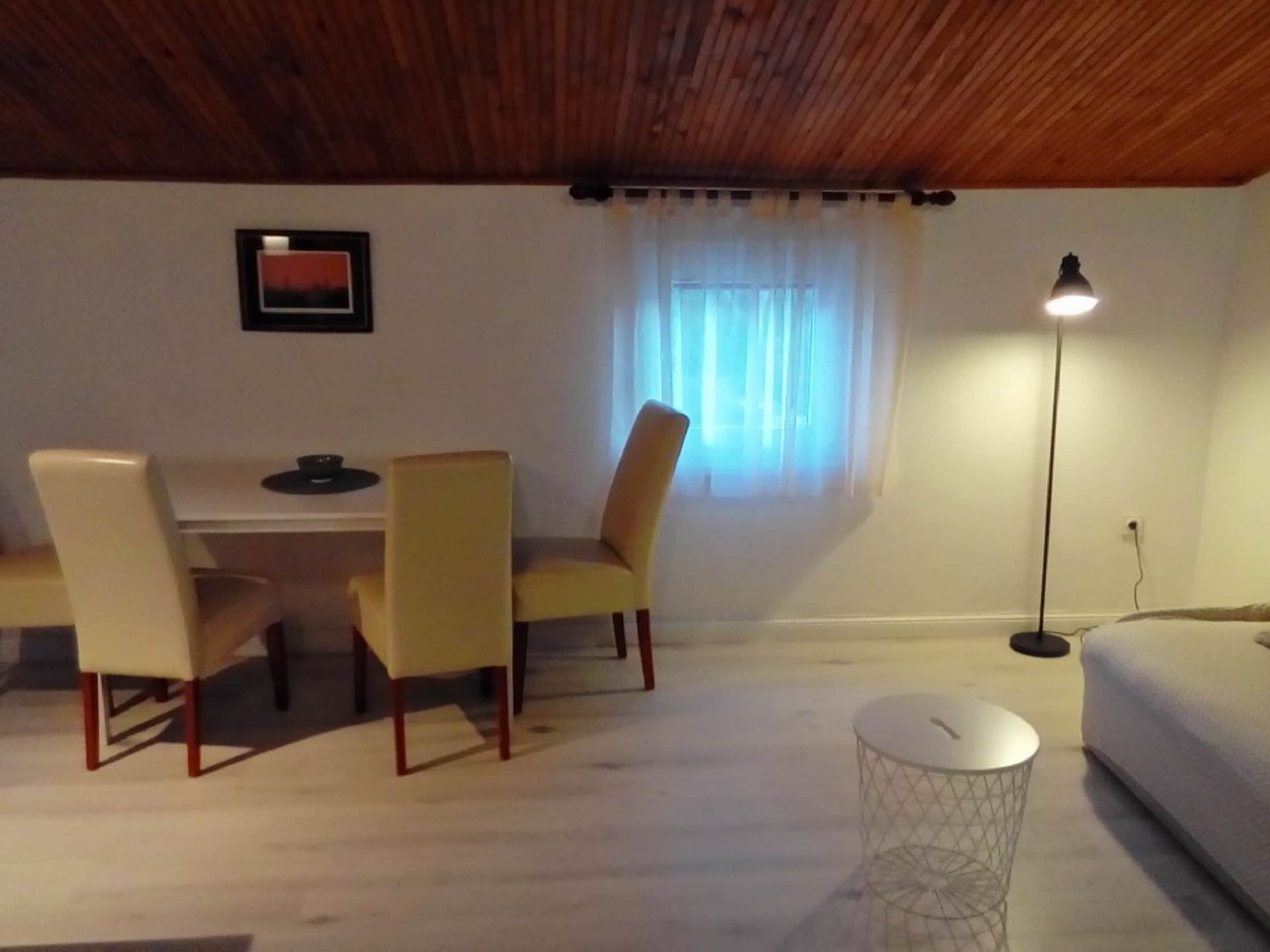 A cozy room with a wooden ceiling, a dining table with chairs, a small round table, a floor lamp, and a bed.